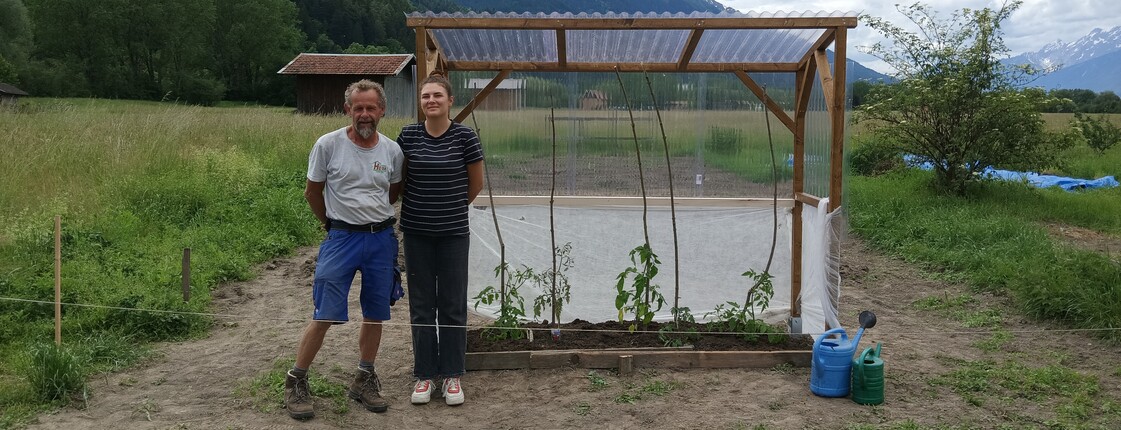 Zwei Mitarbeitende stehen neben einem kleinen, provisorischen Gewächshaus auf einem Feld.