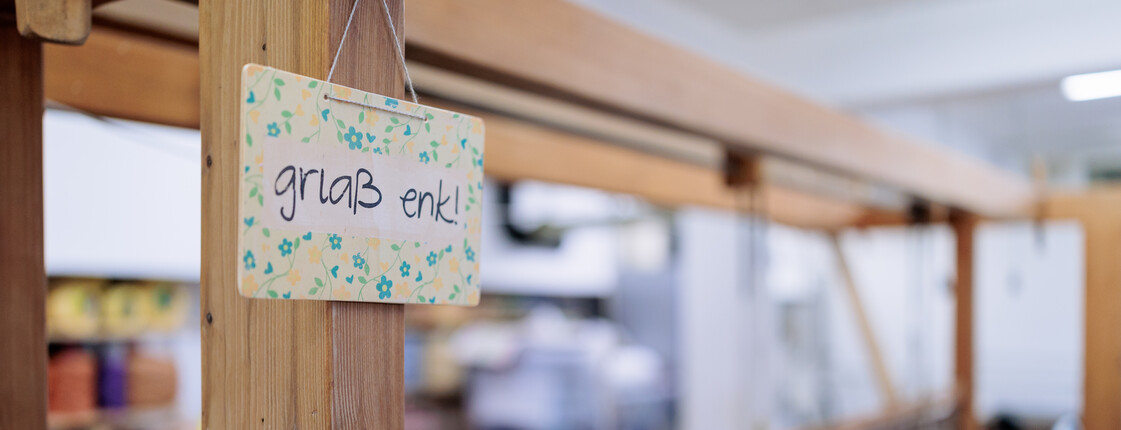 Öffnungszeiten An einem großen Webstuhl aus Holz hängt ein buntes Schild mit der Aufschrift "griaß enk!".