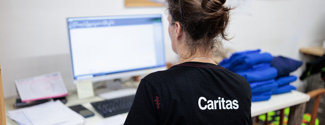 Leitbild des abrakadabra Eine Frau, die ein schwarzes T-Shirt mit weißem Caritas Logo trägt, sitzt an einem Schreibtisch vor einem Bildschirm.